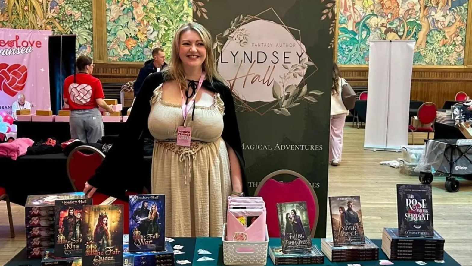 Lyndsey Hall behind a table stacked with her books, in front of a roller banner with her logo, at a book signing event in Swansea.