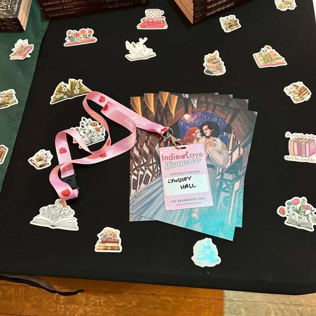 An Indie Love Wales lanyard with Lyndsey Hall's name, on top of a stack of character art and surrounded by bookish stickers.
