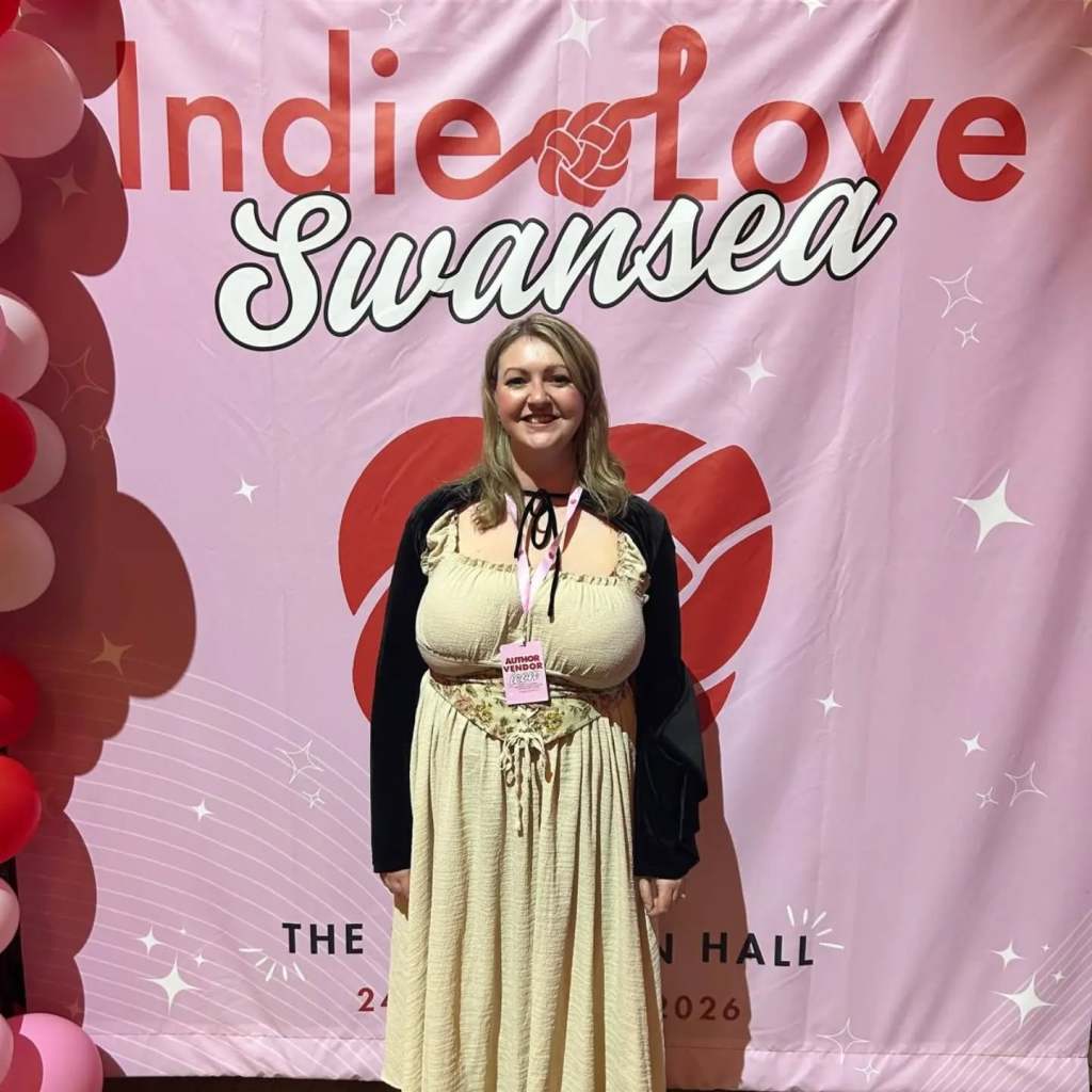 Lyndsey Hall standing in front of a huge pink Indie Love Wales Swansea 2026 sign, wearing a corset dress and hooded capelet.
