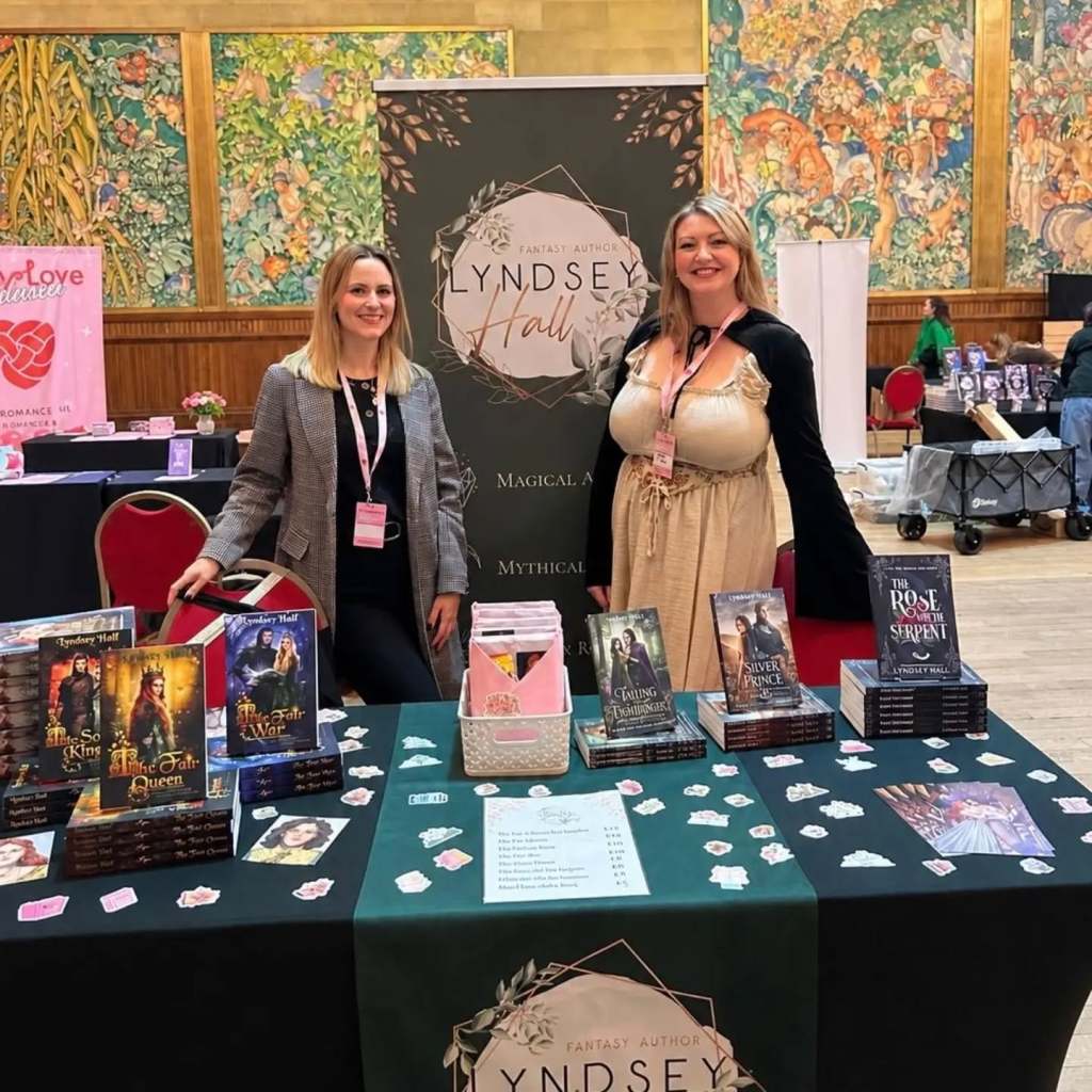 Lyndsey Hall and friend standing behind a table stacked with books at a book signing event in Brangwyn Hall, Swansea.